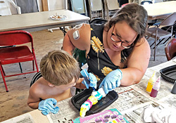 A woman in gloves helping a child squeeze dye onto a folded shirt at a work table