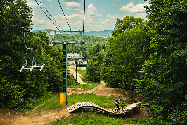 A rider crossing a wooden ladder feature at Burke Mountain Bike Park, with the chairlift and the mountains beyond