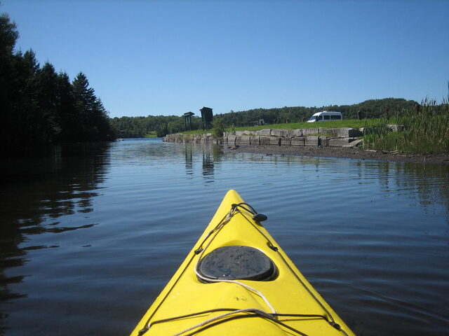 The bow of a yellow kayak on flat water at dusk, with a wooded shoreline ahead