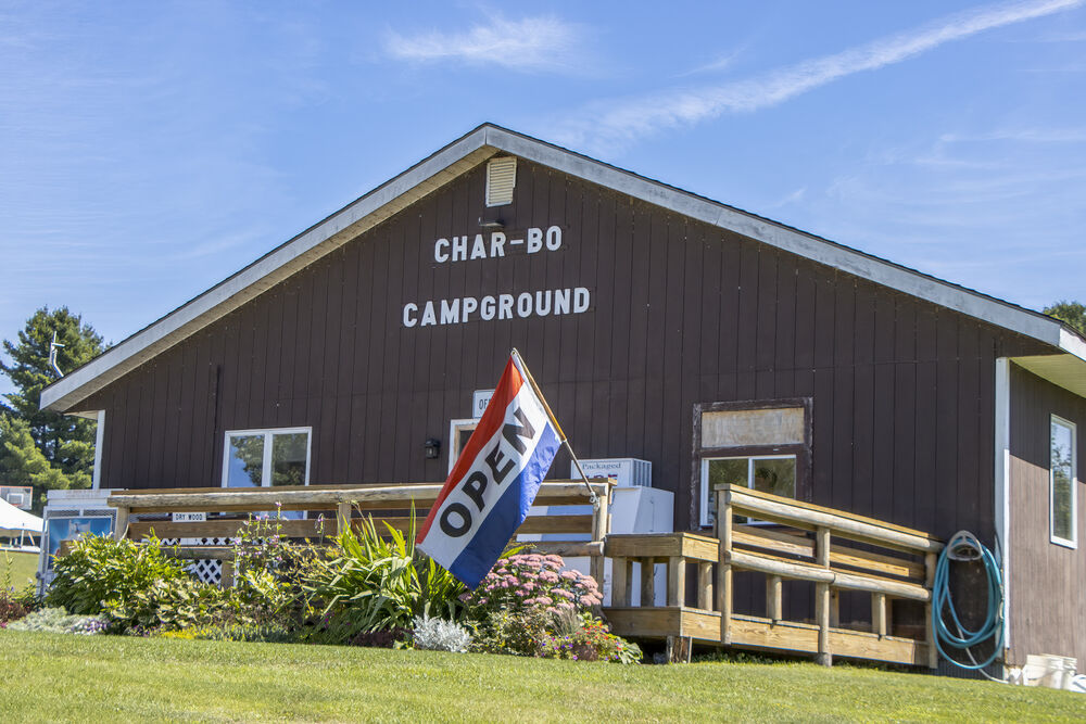 The brown board-and-batten camp store and rec hall with CHAR-BO CAMPGROUND lettering on the gable and an OPEN flag by the steps