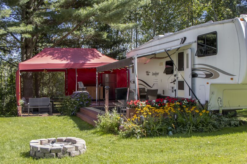 A fifth-wheel beside a red canopy, flower beds and a stone fire ring
