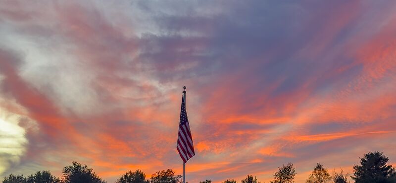 The flag silhouetted against a pink and orange sunset over the treeline