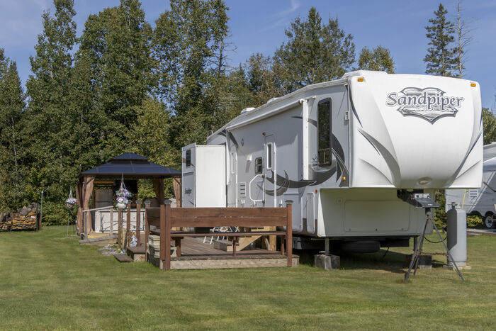 A fifth-wheel beside a gazebo and a railed wooden deck at the edge of the trees