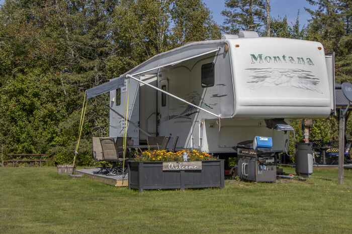 A fifth-wheel with its awning out, camp chairs and a planter of yellow flowers