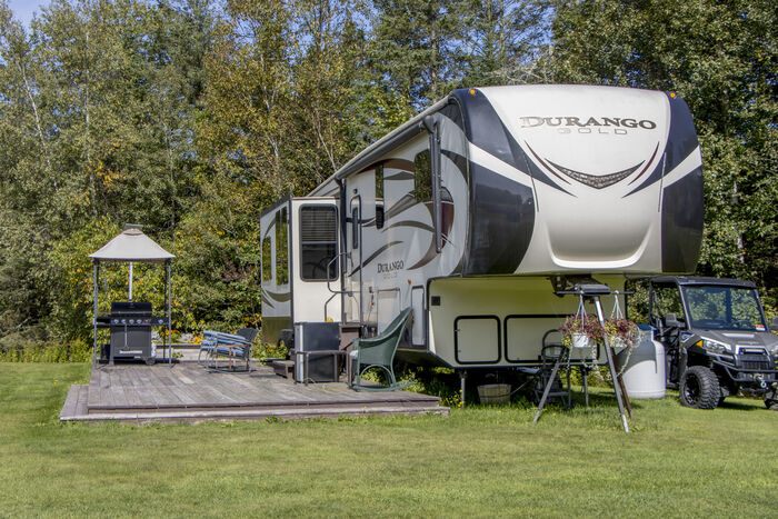 A fifth-wheel with a long wooden deck, a gazebo and a grill under the trees