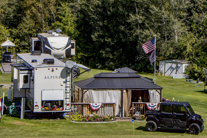 A fifth-wheel with a hardtop gazebo, a flagpole and a side-by-side parked alongside