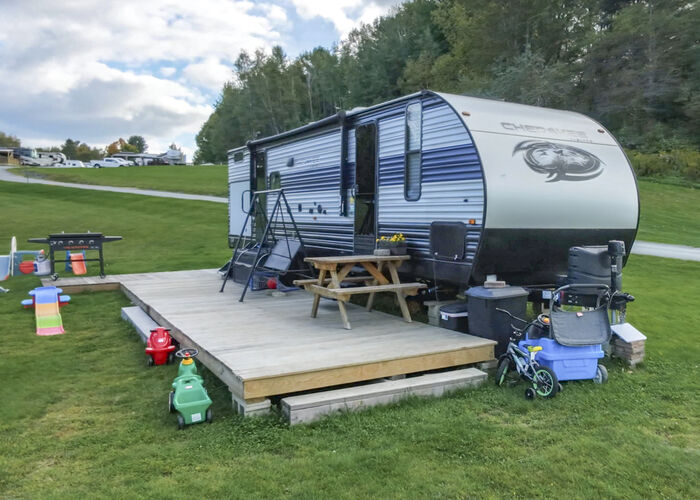 A travel trailer with a wide wooden deck, a picnic table, a grill and children's ride-on toys
