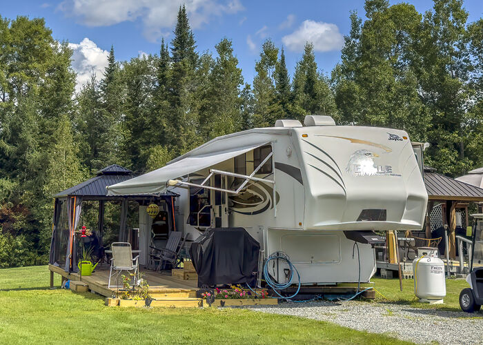 A fifth-wheel with a screened gazebo on a wooden deck, backed by tall spruce